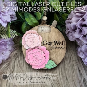 以下が含まれることがあります： ピンクの花柄が描かれた丸い木製オーナメント。"Get Well Soon"と"May"の文字が書かれています。