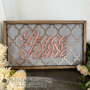 Può includere: Un cartello di legno con uno sfondo geometrico grigio e oro. Le parole "Peace Love Home" sono tagliate nel legno e dipinte di color oro rosa.