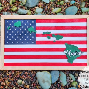 Puede incluir: Una bandera estadounidense enmarcada en madera con un contorno verde del estado de Hawái y la palabra "Hawái" en letras blancas. La bandera está sobre un fondo de pequeñas piedras de colores.