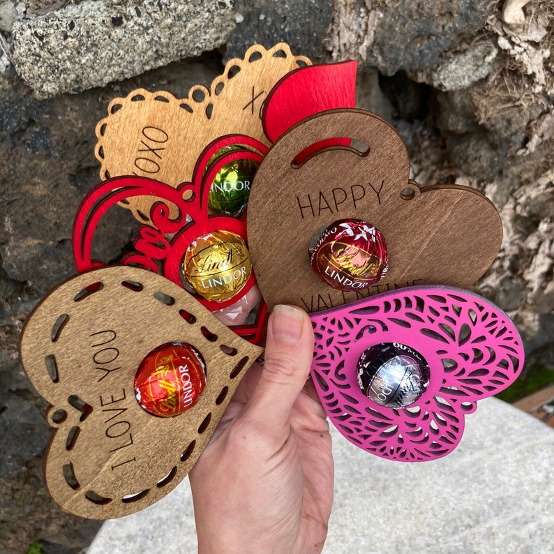 May include: Five wooden heart-shaped holders with intricate designs, each holding a Lindor chocolate truffle. The holders are in various colors, including brown, red, and pink. The holders are engraved with different messages, including "I love you", "Happy Valentine", and "XOXO".
