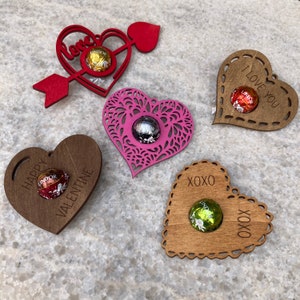 May include: Five wooden heart-shaped ornaments with different designs and sayings. Each heart has a small indentation to hold a chocolate candy. The sayings include "Love", "Happy Valentine", "I Love You", and "XOXO".