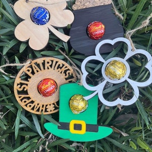 May include: Five wooden ornaments with a St. Patrick's Day theme. Each ornament has a different design, including a shamrock, a pot of gold, a leprechaun hat, and a circle with the text "Happy St. Patrick's Day". Each ornament has a small hole to insert a chocolate candy.