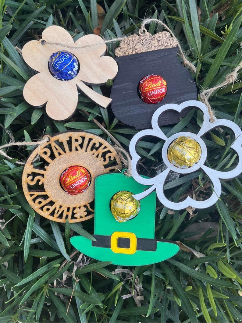 May include: Five wooden ornaments with a St. Patrick's Day theme. Each ornament has a different design, including a shamrock, a pot of gold, a leprechaun hat, and a circle with the text "Happy St. Patrick's Day". Each ornament has a small hole to insert a chocolate candy.
