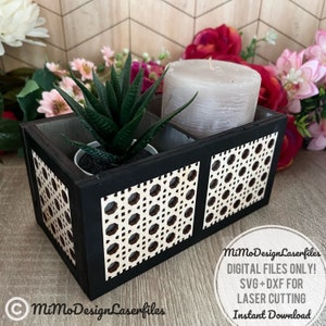 May include: A black wooden planter box with a geometric pattern on the front. The planter has two compartments, one for a small succulent plant and the other for a candle. The planter is sitting on a light brown surface.
