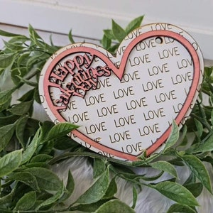 May include: A wooden heart-shaped decoration with a pink outline and the words "Happy Valentine's Day" cut out. The background is white with the word "LOVE" repeated. The decoration is surrounded by green leaves.