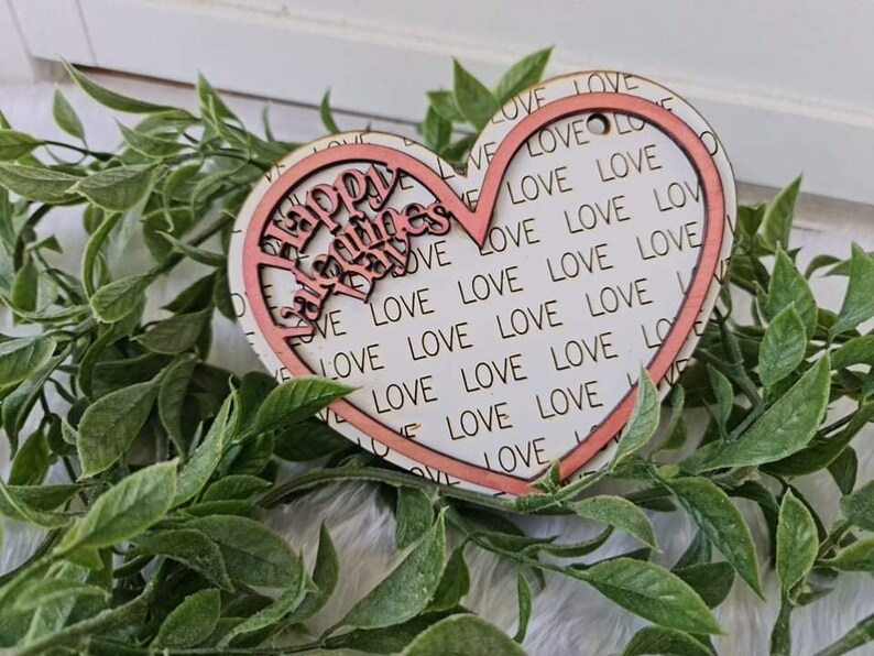 May include: A wooden heart-shaped decoration with a pink outline and the words "Happy Valentine's Day" cut out. The background is white with the word "LOVE" repeated. The decoration is surrounded by green leaves.