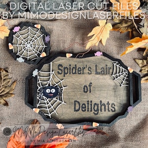 Può includere: Un vassoio di legno con un design di ragno nero e ragnatela. Il vassoio ha un manico ritagliato su ogni lato e dice "Spider's Lair of Delights".
