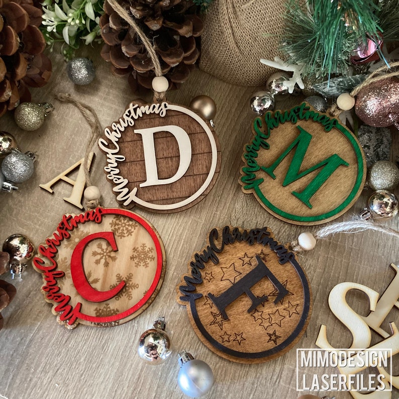 Op de afbeelding: Vier houten kerstversieringen met de letters A, C, D en F in het midden. Elke versiering heeft de woorden "Merry Christmas" rond de rand geschreven. De versieringen liggen op een houten oppervlak met dennenappels en andere kerstversieringen.