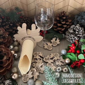 Puede incluir: Anillos de servilleta de reno de madera y encantos para copas de vino con frases de temática navideña. El anillo de servilleta dice "Feliz Navidad". Los encantos tienen frases como "Dashing Through Merlot" y "Es la temporada de ser alegre".