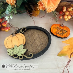 May include: Two wooden pumpkin ornaments with a chevron patterned wooden oval. The larger pumpkin has a leaf and vine design. The smaller pumpkin has the text "Happy Fall" engraved on it.