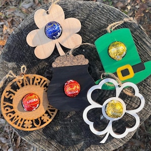 May include: Five wooden ornaments with cutouts in the shape of a shamrock, a pot of gold, a leprechaun hat, a circle with "Happy St. Patrick's Day" text, and a four-leaf clover. Each ornament has a small hole to insert a chocolate candy. The candies are wrapped in gold, blue, and red foil.