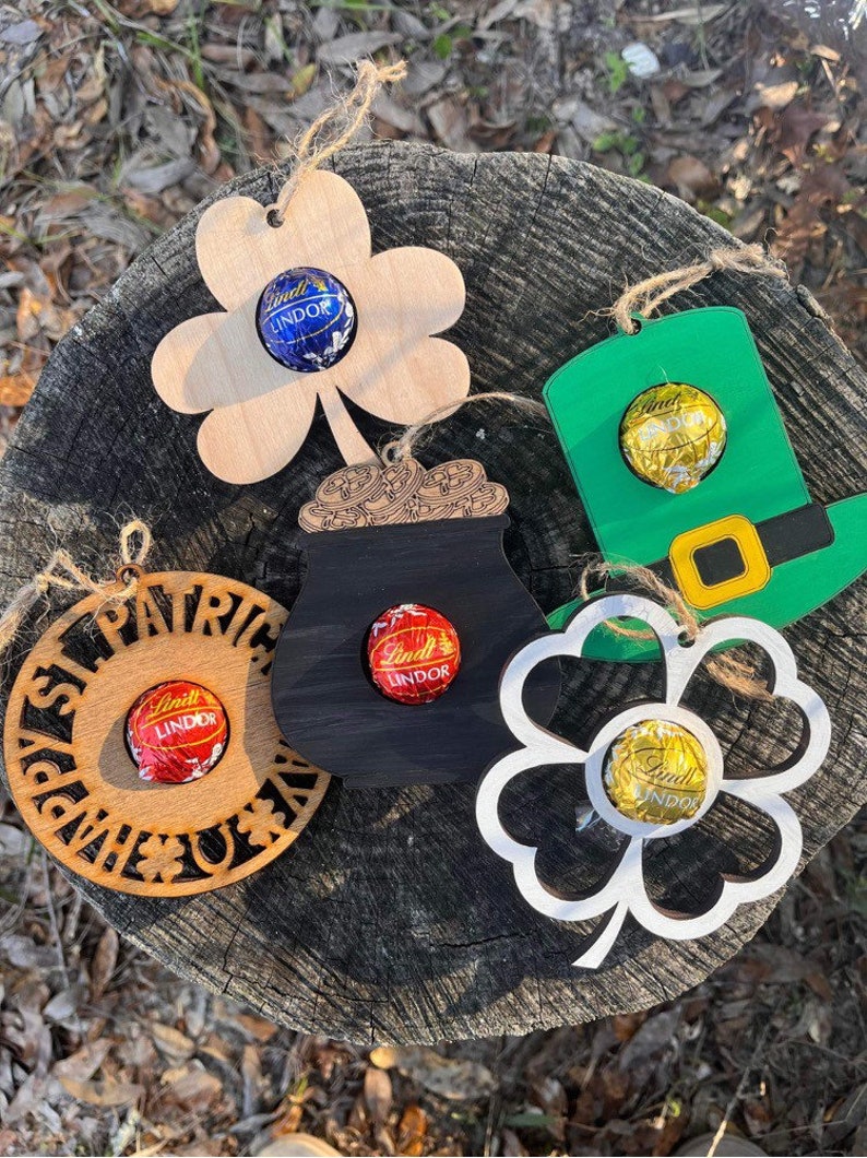 May include: Five wooden ornaments with cutouts in the shape of a shamrock, a pot of gold, a leprechaun hat, a circle with "Happy St. Patrick's Day" text, and a four-leaf clover. Each ornament has a small hole to insert a chocolate candy. The candies are wrapped in gold, blue, and red foil.
