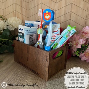 May include: A brown wooden box with a handle, filled with various colorful toothbrushes and toothpaste tubes. The box is labeled "MiMo Design Laserfiles" and the text "DIGITAL FILES ONLY! SVG+DXF FOR LASER CUTTING Instant Download" is visible below the box.