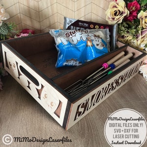 May include: A wooden tray with a cutout design of a marshmallow and the words "How to Delicious S'mores Station". The tray has a compartment for holding skewers and another compartment for holding marshmallows. The tray is brown with a light wood finish.