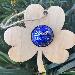 May include: A wooden shamrock cutout with a Lindt Lindor chocolate truffle in the center. The shamrock is light brown and has a hole at the top for hanging.
