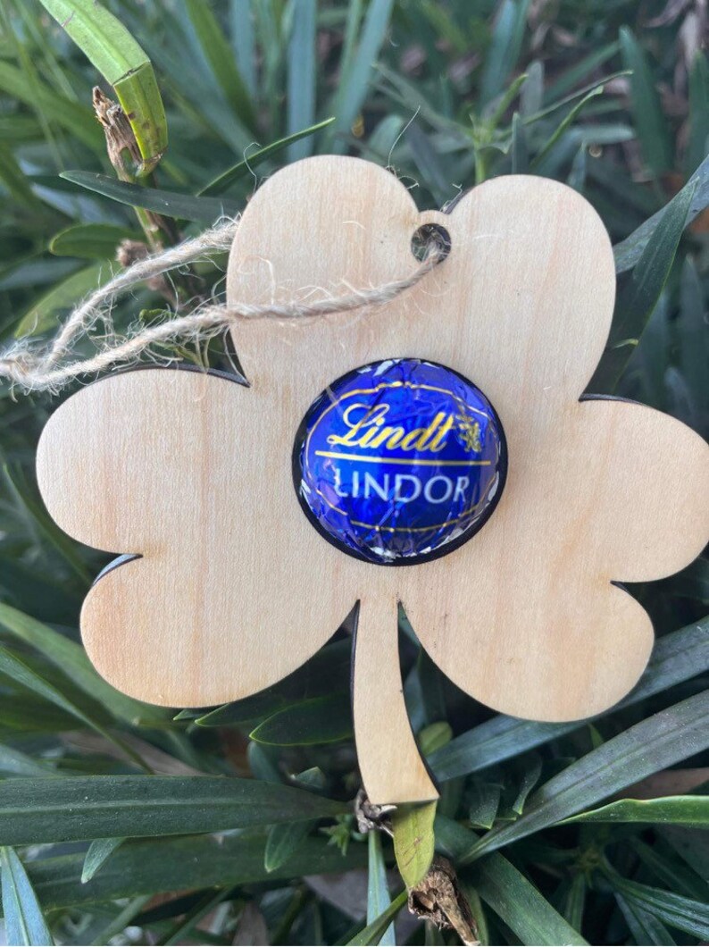 May include: A wooden shamrock cutout with a Lindt Lindor chocolate truffle in the center. The shamrock is light brown and has a hole at the top for hanging.