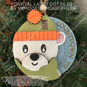 Può includere: Un ornamento natalizio in legno con un orso polare che indossa un cappello verde e arancione. L'ornamento è rotondo e ha il testo "Countdown to Christmas".