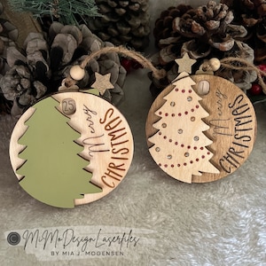 Può includere: Due decorazioni natalizie in legno con un design ad albero. Un ornamento è verde e dice "Buon Natale" con il numero 25. L'altro ornamento è marrone e dice "Buon Natale" con il numero 1.