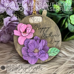 Può includere: Un ornamento rotondo in legno con un design tagliato al laser che presenta due fiori, uno rosa e uno viola, con foglie verdi. L'ornamento ha il testo "You Happy Mother's Day" inciso.