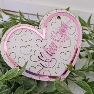 May include: A heart-shaped ornament with a pink border and the words "I love you" in cursive. The ornament has a white background with a pattern of small hearts. A small hole is at the top for hanging.