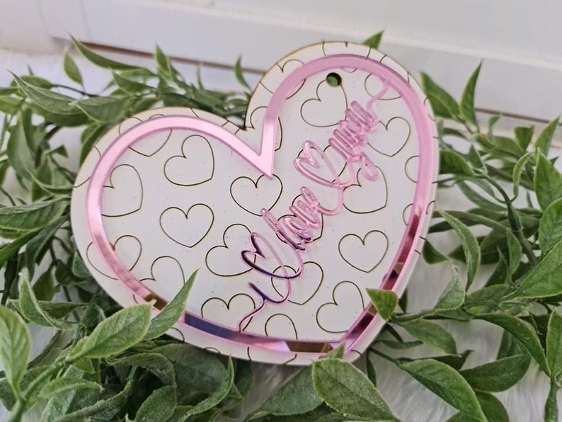 May include: A heart-shaped ornament with a pink border and the words "I love you" in cursive. The ornament has a white background with a pattern of small hearts. A small hole is at the top for hanging.