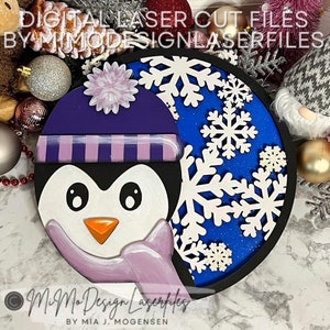 May include: A round wooden sign with a blue background and white snowflakes. A penguin wearing a purple hat with a pink and white striped band and a pink scarf is on the left side of the sign. The text "MiMo Design Laserfiles" is at the bottom of the sign.