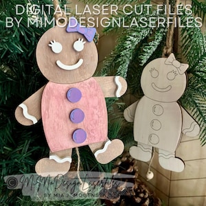 May include: Two wooden gingerbread people ornaments, one with a pink dress and purple buttons, the other in natural wood. Both have white icing details and are hanging from a Christmas tree.