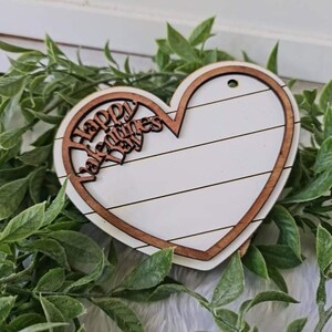 May include: A wooden heart-shaped decoration with a white center and a brown border. The words "Happy Valentines Day" are cut out of the top portion. The heart is surrounded by green leaves.