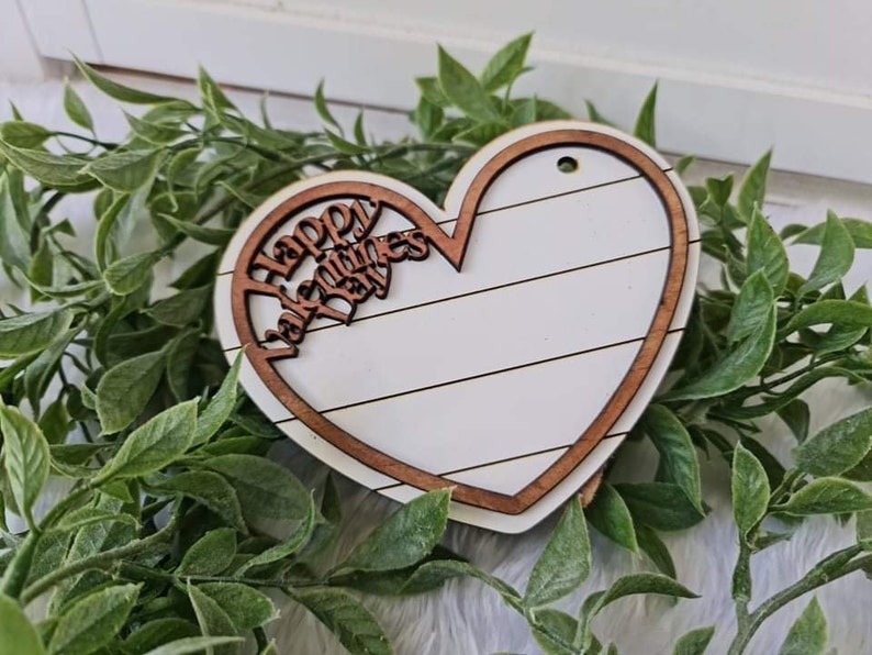 May include: A wooden heart-shaped decoration with a white center and a brown border. The words "Happy Valentines Day" are cut out of the top portion. The heart is surrounded by green leaves.
