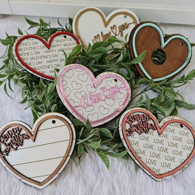 May include: A collection of heart-shaped wooden ornaments with various designs and text. Some hearts have the words "Happy Valentine's Day" or "I love you". The ornaments are in shades of brown, white, pink, and red, and are arranged on a bed of green leaves.