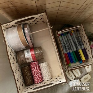 May include: Two wooden boxes with cut-out designs. The larger box contains rolls of ribbon, twine, and string in various colors and textures. The smaller box contains a set of colorful Sharpie markers. The boxes have labels that read "Tools", "Keychains", "Ink/Paint", "Bottle", and "Spl", likely for organizing craft supplies.