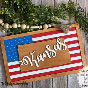 Könnte beinhalten: Ein Holzschild mit einem rot-weiß-blauen amerikanischen Flaggen-Design. Der Bundesstaat Kansas ist aus der Flagge ausgeschnitten und das Wort "Kansas" ist in weiß auf braunem Hintergrund geschrieben.