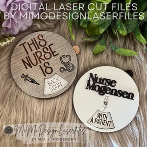 May include: Two round wooden signs with a rotating dial. The top sign is brown with the text "This Nurse Is" and a stethoscope. The bottom sign is white with the text "Nurse Mogensen" and a syringe. Both signs have a rotating dial with the text "Back Soon" and "With a Patient".