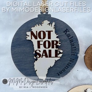 Può includere: Un ornamento rotondo in legno con sfondo blu e un ritaglio della Groenlandia in legno più chiaro. Le parole "NOT FOR SALE" sono ben visibili. Il testo "Kalaallit Nunaat" è curvo lungo il bordo destro. Progettato da MiMoDesignLaserfiles.