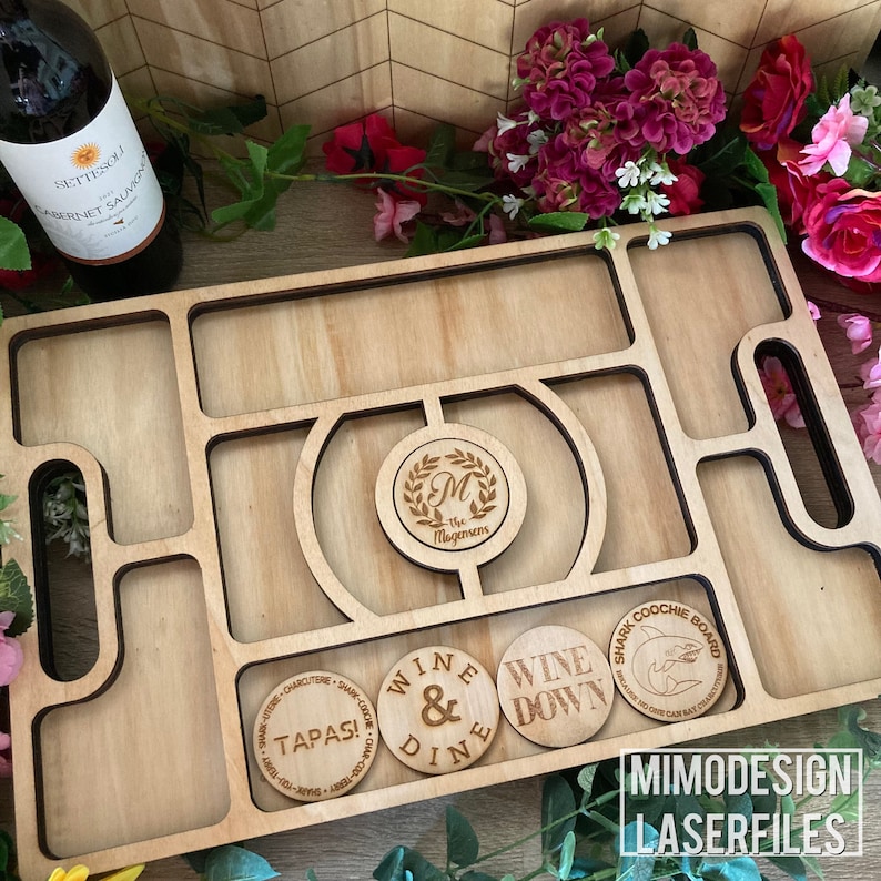 Op de afbeelding: Een houten serveerblad met meerdere compartimenten en uitsparingen. Het blad is voorzien van lasergegraveerde ontwerpen, waaronder een monogram met de initialen "M" en de tekst "The Magensens", evenals de zinnen "Wine & Dine", "Wine Down" en "Tapas!" Het blad bevat ook een haaienvormige uitsparing met de tekst "Shark Coochie Board".