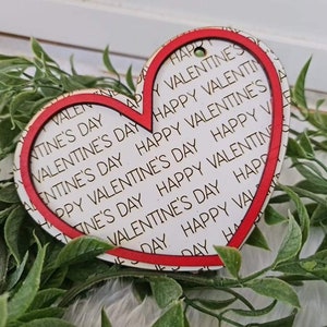 May include: A heart-shaped wooden ornament with a red border and the words "Happy Valentine's Day" printed repeatedly on a white background. The ornament has a small hole at the top for hanging. The ornament is surrounded by green foliage.