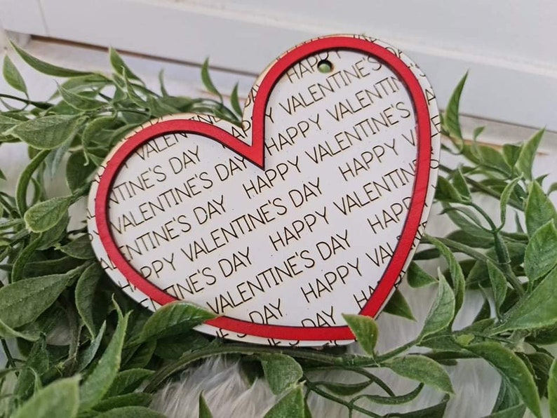 May include: A heart-shaped wooden ornament with a red border and the words "Happy Valentine's Day" printed repeatedly on a white background. The ornament has a small hole at the top for hanging. The ornament is surrounded by green foliage.