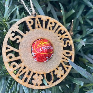 May include: A wooden ornament with the words "Happy St. Patrick's Day" cut out around the edge. A Lindor chocolate is in the center of the ornament.
