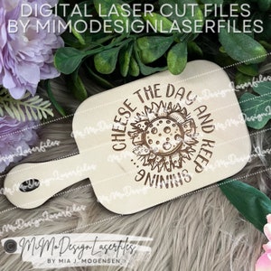 Può includere: Un tagliere in legno con manico, che presenta un design a forma di sole con il testo "Cheese the day and keep shining".
