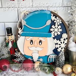 May include: A blue and white wooden nutcracker face with a white beard and mustache. The nutcracker is surrounded by white snowflakes on a dark blue background. The background is a brown wooden circle.
