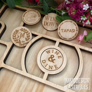 Op de afbeelding: Een set van houten gegraveerde cirkels met verschillende zinnen en ontwerpen. De cirkels zijn gerangschikt op een houten bord met uitsparingen voor elke cirkel. De zinnen omvatten "Shark Coochie Board", "Wine Down", "Tapas!", "Wine & Dine", en "The Mogensene".