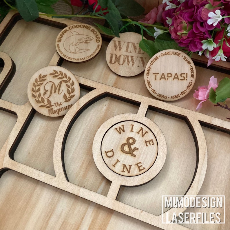 Op de afbeelding: Een set van houten gegraveerde cirkels met verschillende zinnen en ontwerpen. De cirkels zijn gerangschikt op een houten bord met uitsparingen voor elke cirkel. De zinnen omvatten "Shark Coochie Board", "Wine Down", "Tapas!", "Wine & Dine", en "The Mogensene".