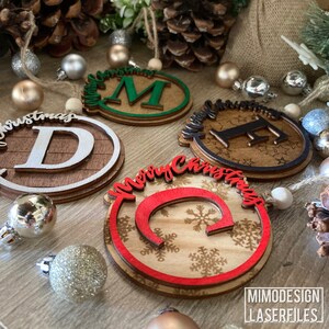 Op de afbeelding: Vier houten kerstversieringen met verschillende ontwerpen. Een versiering is groen met de letter "M" in het midden en de tekst "Merry Christmas". Een andere versiering is bruin met de letter "F" in het midden en de tekst "Merry Christmas". Een derde versiering is bruin met de letter "D" in het midden en de tekst "Christmas". De vierde versiering is bruin met de letter "C" in het midden en de tekst "Merry Christmas".