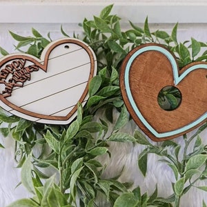 May include: Two wooden heart-shaped ornaments. One is white with brown trim and the words "Happy Valentines Day". The other is brown with a teal border. Both have a hole for hanging. Decorative Valentine's Day decor.