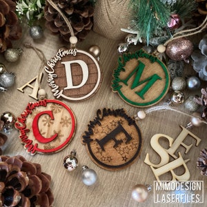 Op de afbeelding: Vijf houten kerstversieringen met de letters A, C, D, E en M in het midden van elke versiering. Elke versiering heeft de tekst "Merry Christmas" rond de rand. De versieringen liggen op een houten oppervlak met dennenappels en kleine decoratieve ballen.