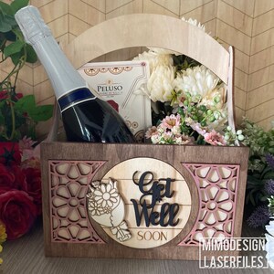 Könnte beinhalten: Ein Geschenkkorb aus Holz mit einem Griff, verziert mit einem rosa-weißen Blumendesign. Der Korb enthält eine Flasche Sekt, eine Schachtel Kekse und einen Blumenstrauß. Der Korb hat ein Schild mit der Aufschrift "Get Well Soon".