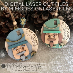 May include: Two wooden nutcracker ornaments with a countdown to Christmas design. Each ornament features a blue and green painted nutcracker face with a number 25 on the side. The ornaments are on a brown and white fuzzy surface.