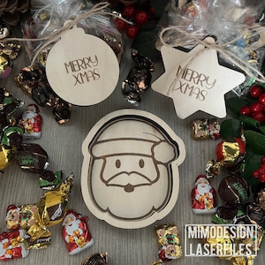 Può includere: Addobbi natalizi in legno con il testo "Merry Xmas". Gli addobbi hanno la forma di una stella, un cerchio e una testa di Babbo Natale. Nell'immagine ci sono anche alcuni cioccolatini.