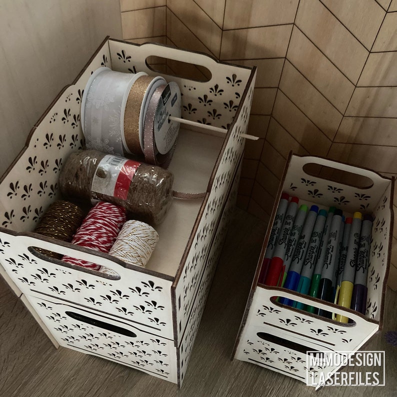 May include: Two wooden storage boxes with intricate cut-out designs. The top box is filled with spools of ribbon and twine in various colors, while the bottom box holds a stack of colorful markers.