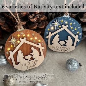 Può includere: Due ornamenti natalizi in legno con un ritaglio della scena della Natività. L'ornamento a sinistra è marrone con glitter dorato e il testo "JOY TO THE World". L'ornamento a destra è blu con glitter argentato e il testo "O HOLY NIGHT Silent Night".